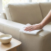 A hand with a wedding band wipes a light faux leather sofa with a white cloth. A spray bottle, foamy solution, and brush sit on a side table.