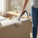 Vue en contre-plongée (buste uniquement) d’une personne nettoyant à la vapeur un canapé en tissu clair. La vapeur visible suggère un nettoyage en profondeur et écologique, dans une pièce lumineuse.