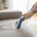 Gloved hands operate a modern sofa cleaner on a light grey fabric sofa, showing foam. Bright, clean living room setting.