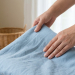 Close-up of hands gently folding a pale blue linen fabric. A blurry wicker basket and bright window are in the serene background.
