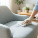 Main gantée d’une personne nettoyant délicatement une tache sur une chaise en tissu gris clair à l’aide d’un spray et d’un chiffon en microfibre, dans un salon moderne et lumineux.