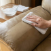Close-up of a hand gently cleaning a beige leather sofa with a white cloth, revealing a clean, lustrous section next to a soiled area. A bowl of soapy water is nearby under soft natural light.