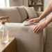 Hands gently clean a beige microfiber sofa with a white cloth, a spray bottle on a wooden side table. Modern, well-lit living room.