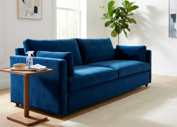 A deep sapphire blue velvet sofa in a bright modern living room. A side table holds a cleaning brush, microfiber cloth, and spray bottle.