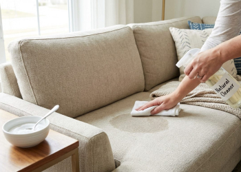 A hand cleans a damp spot on a beige sofa with a "Natural Cleaner" spray bottle and a white cloth. A bowl of baking soda is nearby.