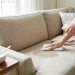 A hand cleans a damp spot on a beige sofa with a "Natural Cleaner" spray bottle and a white cloth. A bowl of baking soda is nearby.