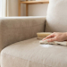 A hand applies white baking soda with a microfiber cloth onto a light Alcantara sofa, with a brush nearby, in a bright living room.