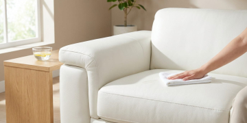 Hand cleaning a white leather sofa with a cloth. Lemon water on a wooden table. Bright room with natural light and a blurred plant.